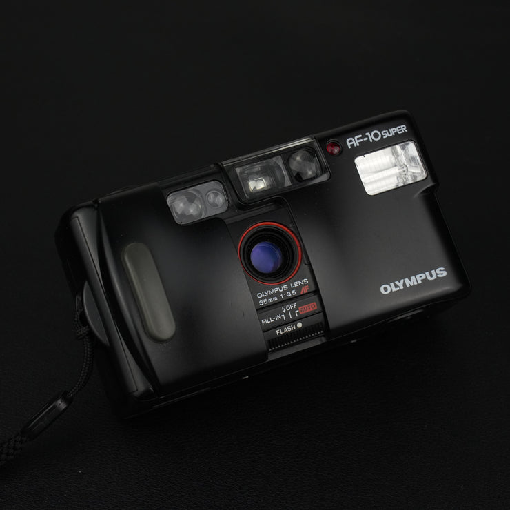 Dettaglio della fotocamera Olympus AF-10 Super con sportellino anteriore aperto che rivela l’obiettivo 35mm f/3.5.